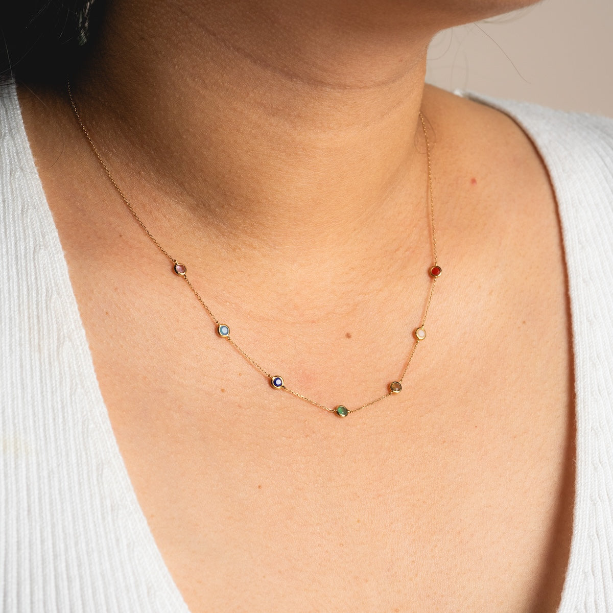 A close-up of someone wearing the Solid Gold Chakra Necklace Sample, crafted in 9k yellow gold with natural multicolored gemstones. Paired with a white V-neck, the warm skin and neutral background highlight the necklaces vibrant charms.
