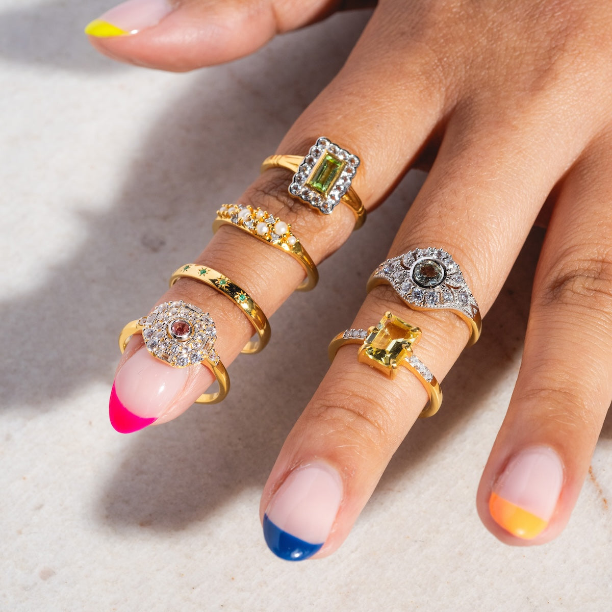 A hand with colorful French tip nails wears six gold rings, including the Madeline Pearl and Diamond Scatter Ring, plus vintage-inspired rings with rectangular, round, and star details, all displayed on a light surface.