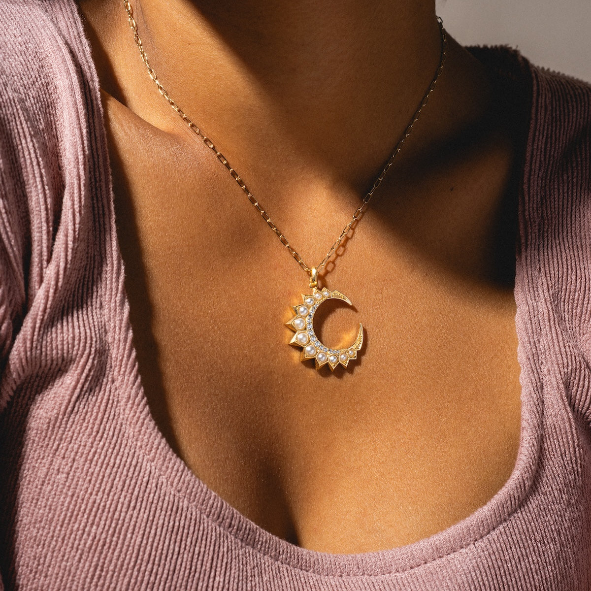 A person wearing the Selene Crescent Necklace Sample, a gold plated crescent moon and sun pendant with cubic zirconia accents, paired with a textured mauve top.