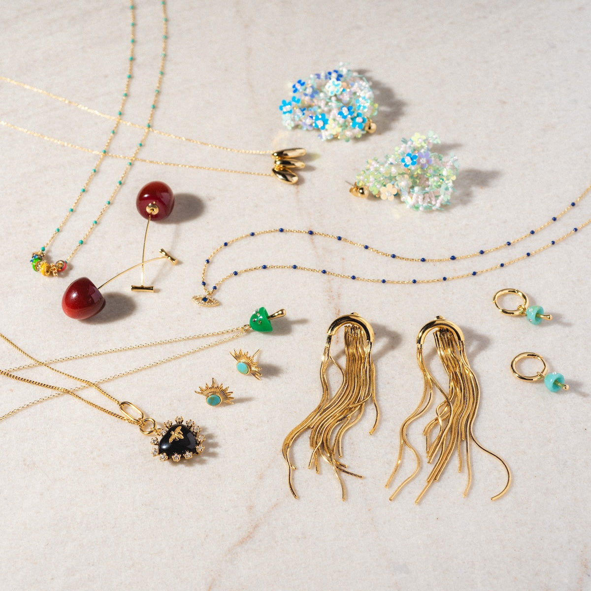Cherry and Stem Earrings displayed on a light marble surface, alongside yellow gold plated necklaces with gemstone pendants, unique long wavy and sparkling floral earrings, plus beaded jewelry and chic green or cherry earring styles.