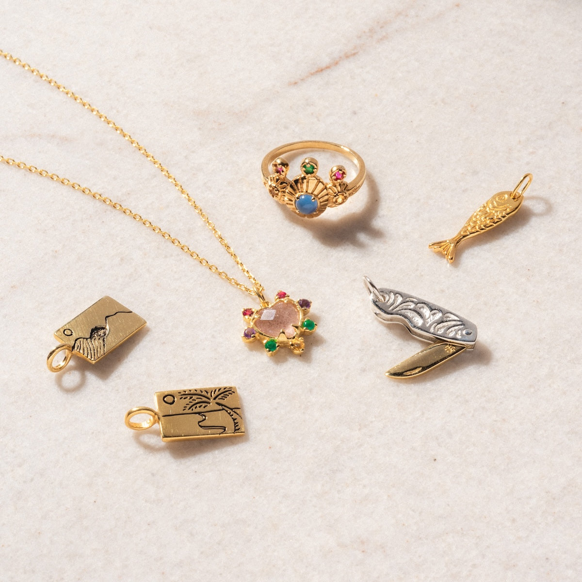 Assorted jewelry on a light surface, featuring the Adore Necklace—an 18k gold vermeil piece with a floral pendant—alongside a gold ring with colorful stones, a fish charm, a silver knife charm, and three engraved rectangular gold charms.