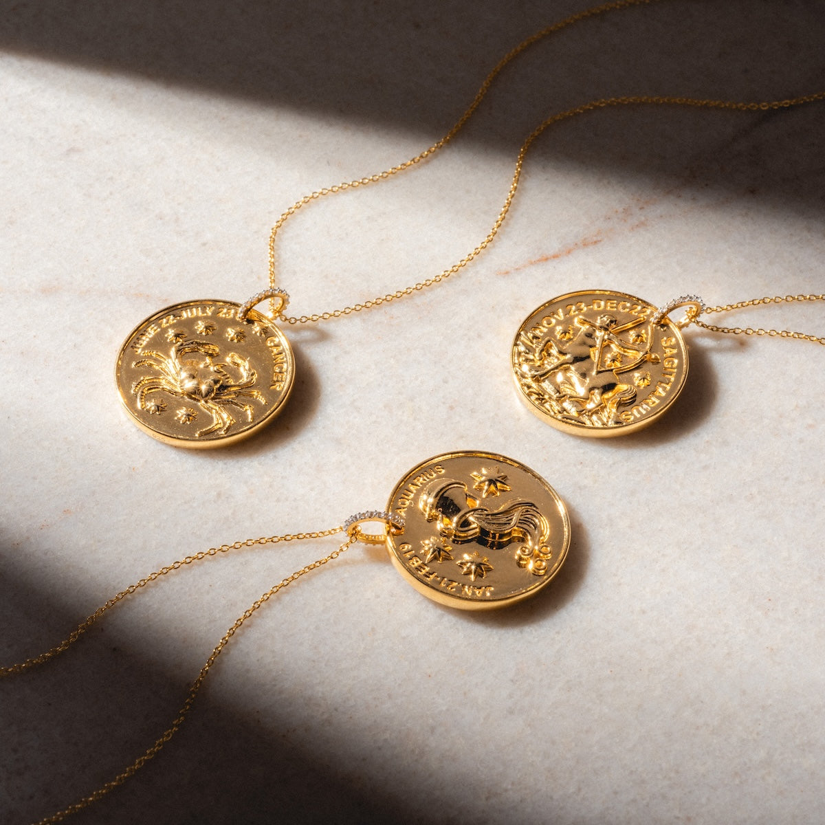 Three Vintage Inspired Zodiac Coin Necklaces with round gold vermeil pendants, engraved with Sagittarius, Capricorn, and Aquarius symbols and dates, are displayed on a light marble surface.