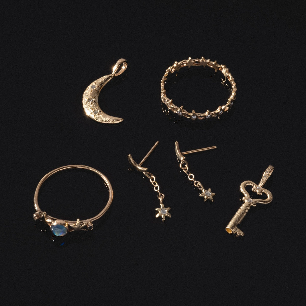 Five pieces of 14k solid yellow gold jewelry on a black background: a crescent moon pendant, a textured ring, a ring with blue stone and small stars, Moon and Stars Drop Earrings, and a key-shaped pendant.