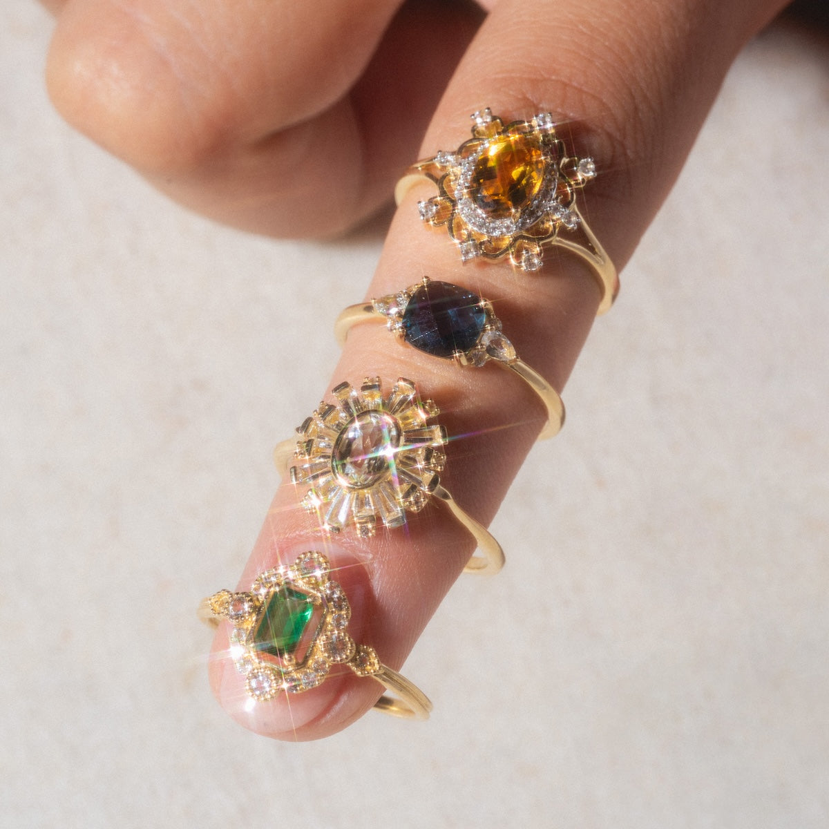 A hand displays the 14k Blue Topaz and White Sapphire Ring, crafted from recycled gold, stacked with other vibrant gemstone rings against a light background.