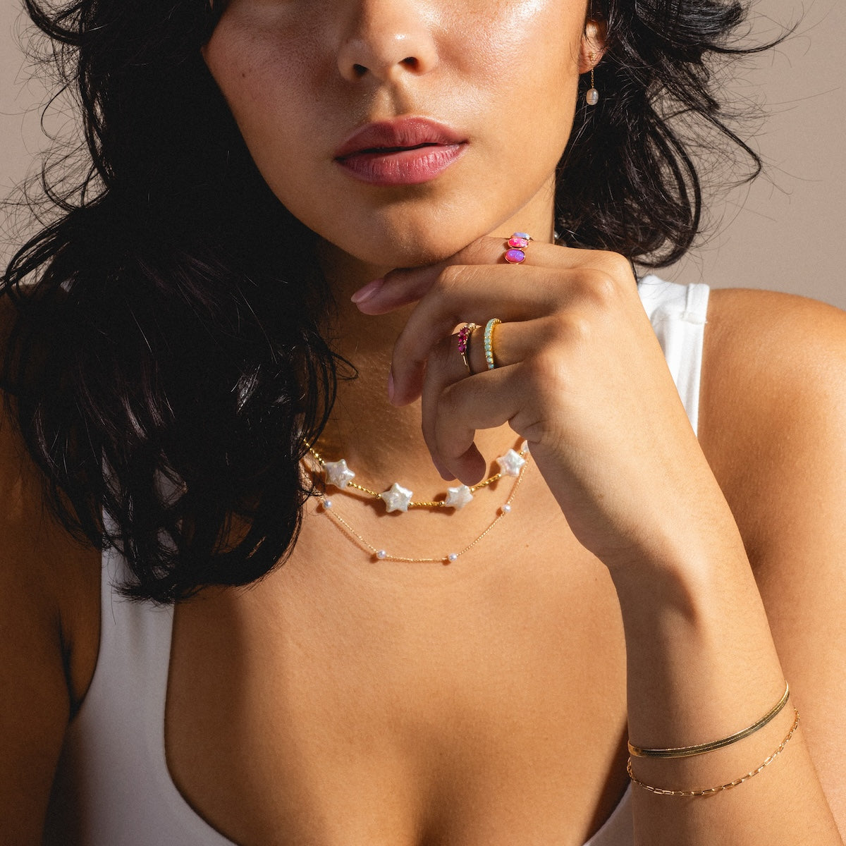 A woman with wavy dark hair, wearing the Slinky Herringbone Bracelet Sample (7 inch) in 18k yellow gold, layered necklaces, rings, and a white tank top, rests her hand near her lips to highlight pink and gold jewelry; her face is partly visible.