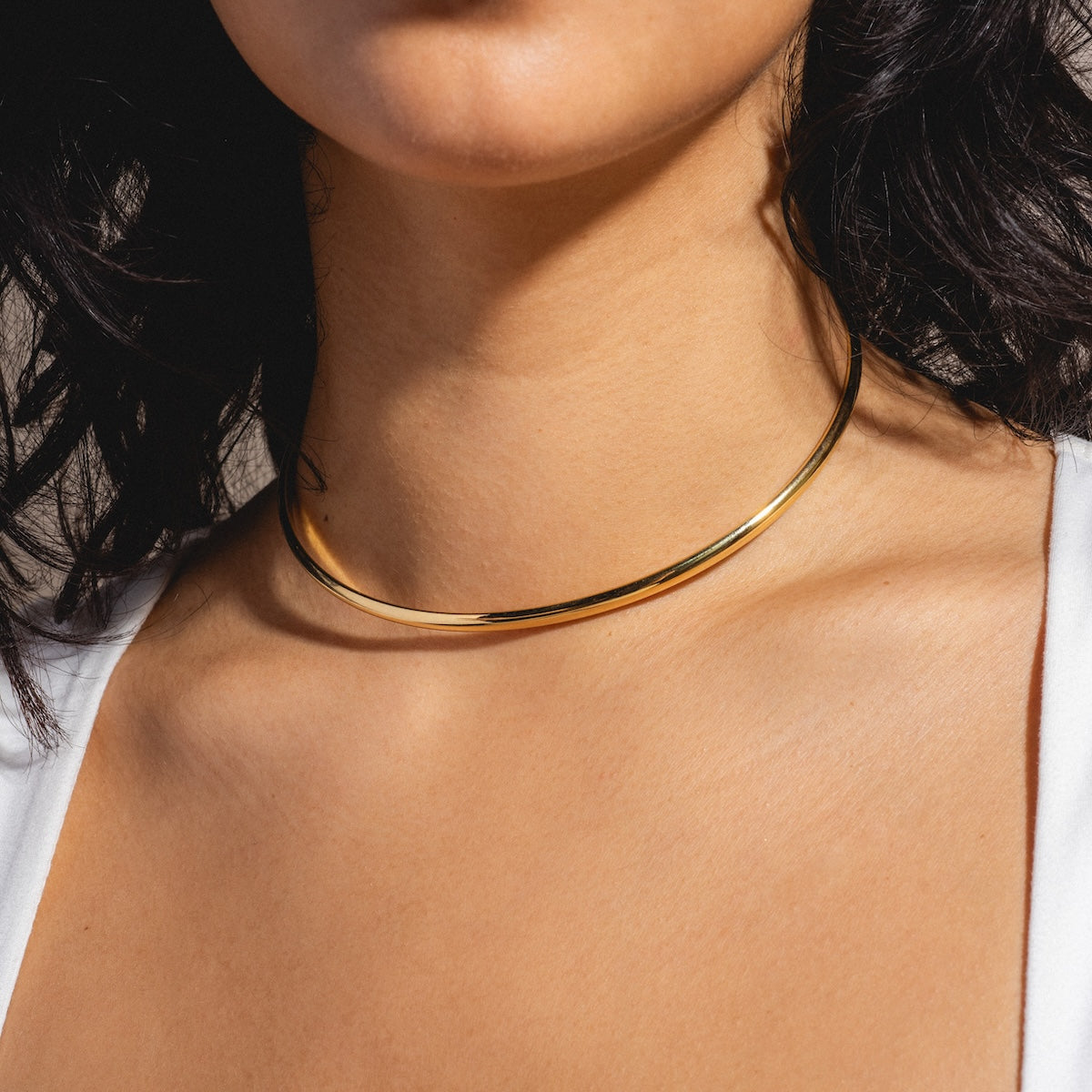 A close-up of a woman wearing the Nikita Cuff Necklace Sample, a 14k yellow gold plated choker, against her bare neck and collarbone, with dark hair and part of a white top visible.