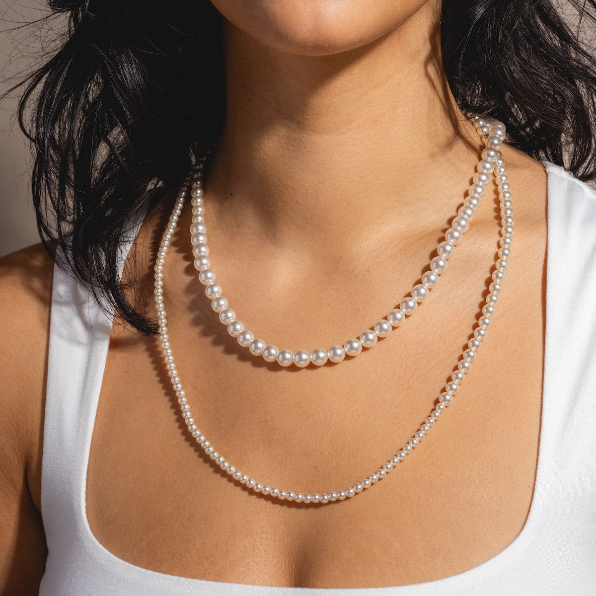 A woman wears the Louise Pearl Necklace, a 14k gold-plated Swarovski pearl piece with large pearls, layered with a long strand of smaller pearls. Only her neck, shoulders, and part of her face are visible as she dons a white top.