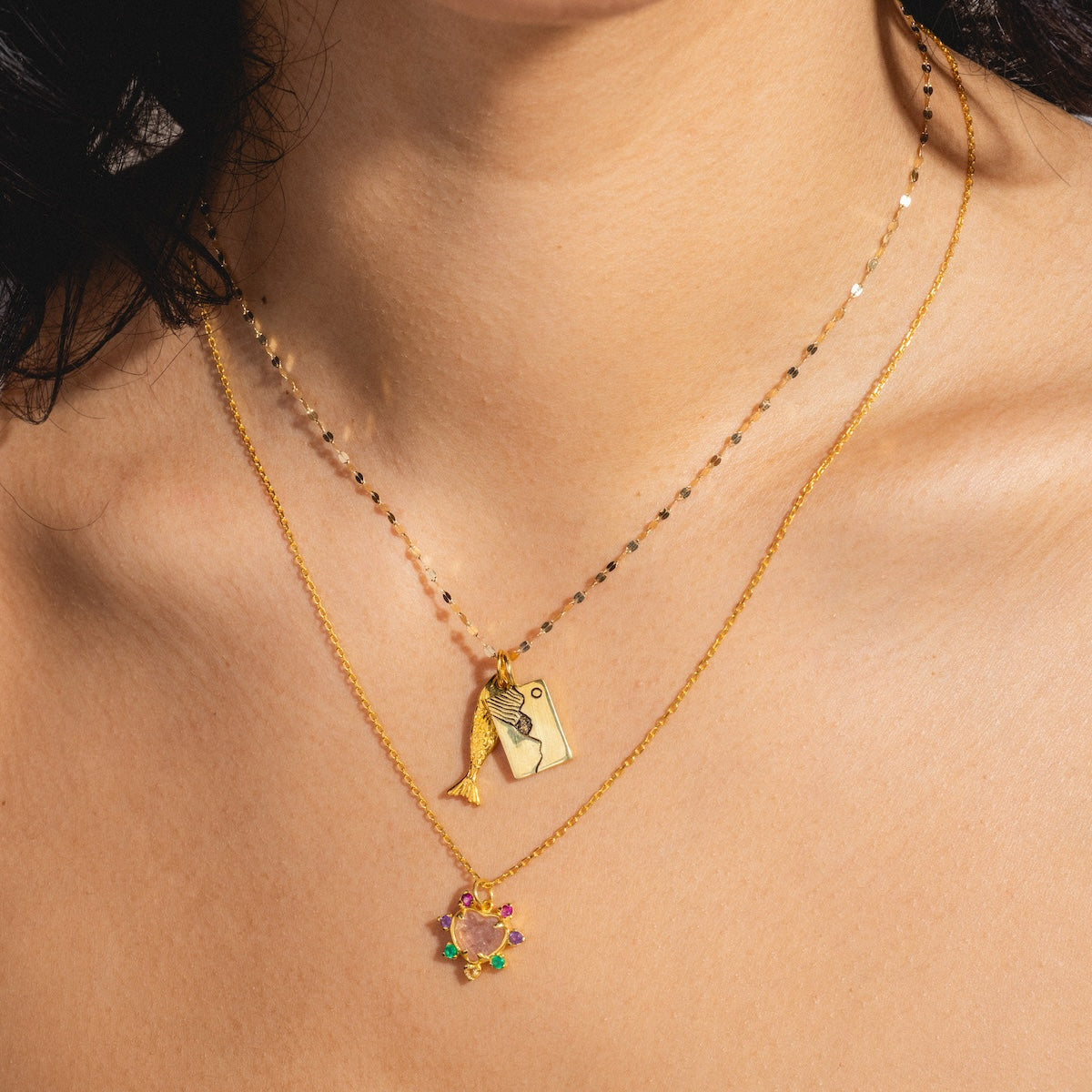 A close-up of a person’s neck wearing two gold vermeil Charms by Hunt of Hounds necklaces: one featuring rectangular and fish charms, the other with a round, colorful gemstone pendant, set against a neutral background.