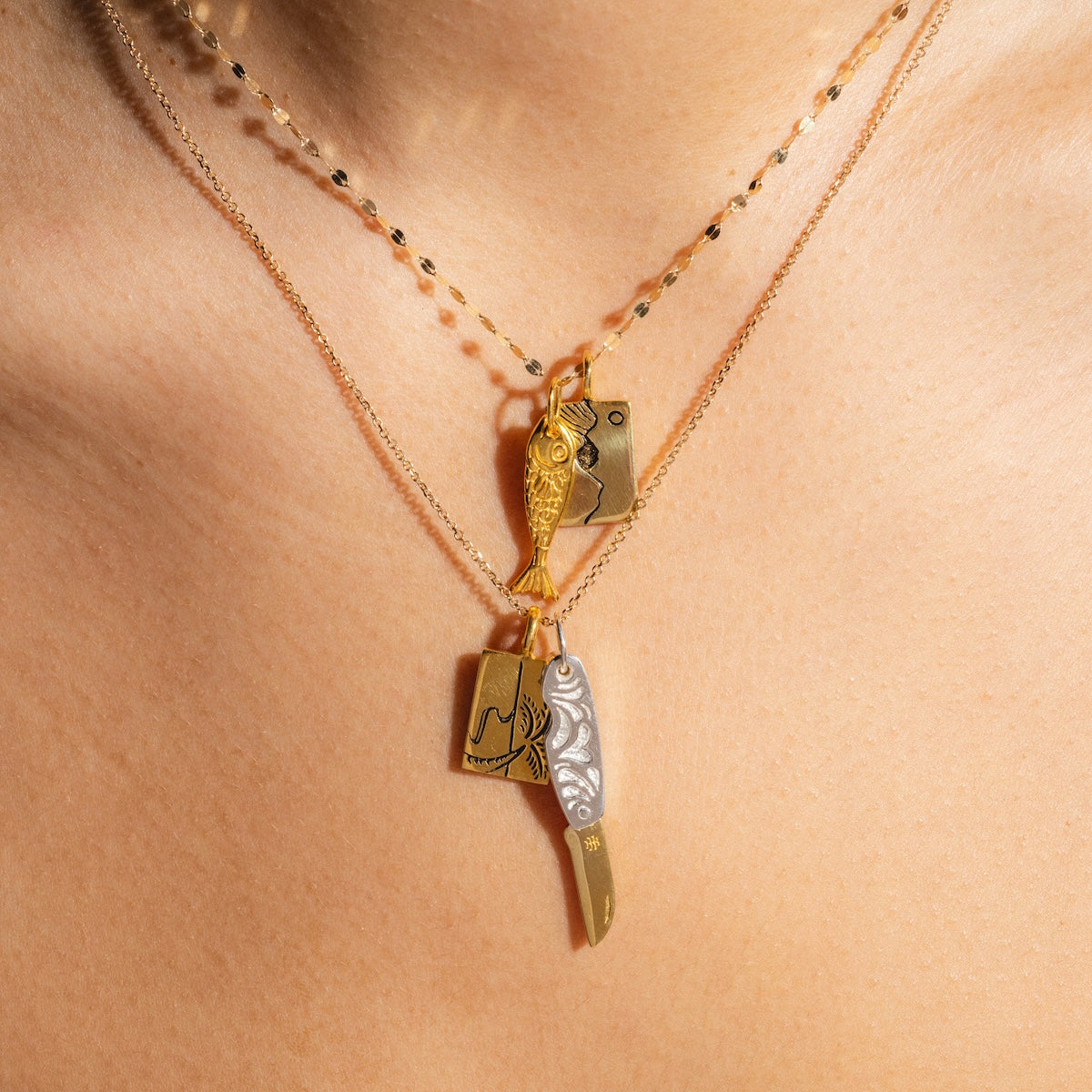 Close-up of Charms by Hunt of Hounds: layered gold vermeil necklaces on skin, with intricate fish, cleaver, and knife-shaped charms.