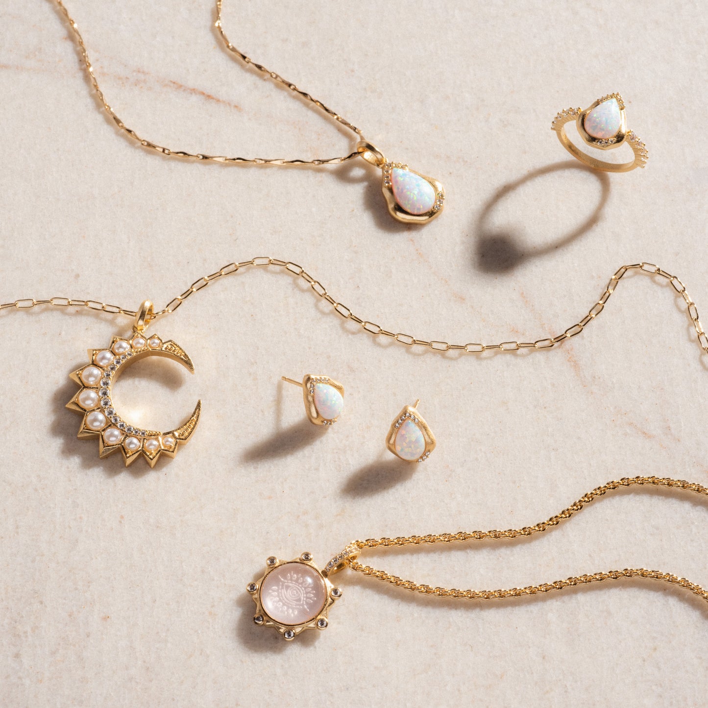 Luster Studs: Teardrop-shaped opal earrings, displayed with two 14k gold plated necklaces, a teardrop opal ring, a pearl crescent moon pendant, and a round pink pendant on a beige stone surface.