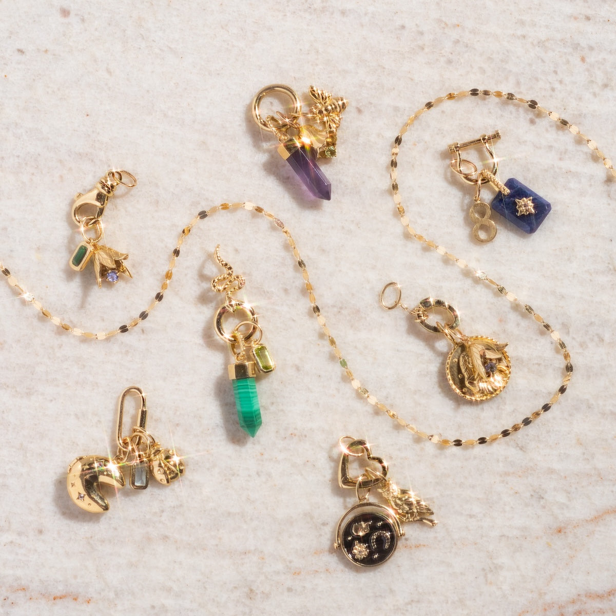 A Solid Gold Charm Clip rests on light marble, surrounded by gold necklaces and intricate charms with colored stones, crystals, moons, hands, and circular pendants—perfect for easy bracelet attachment.