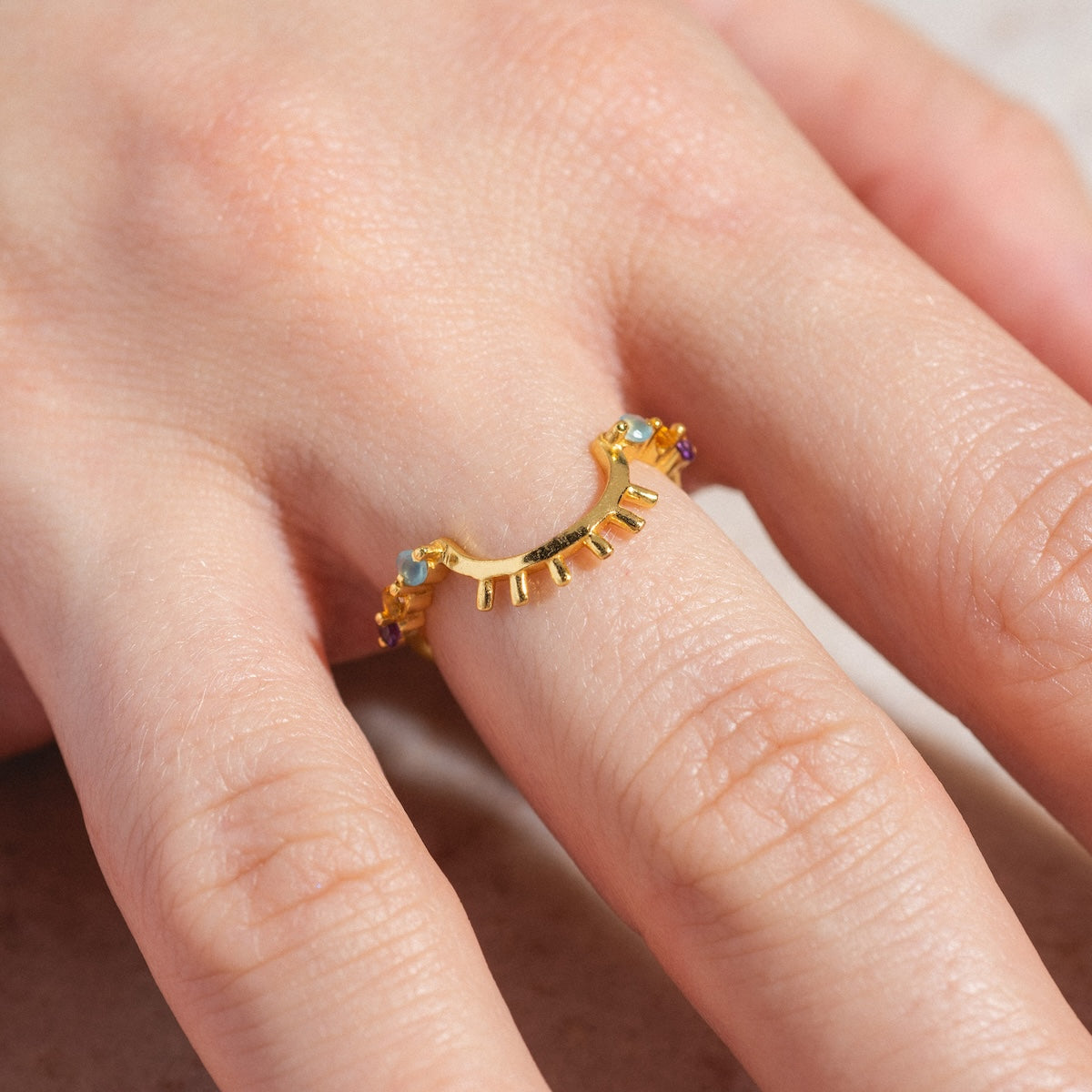 A close-up of a hand wearing the Sunbeam Ring Sample Size 6, an 18k yellow gold plated ring featuring a unique curved design and small amethyst gemstone accents, against a light background.