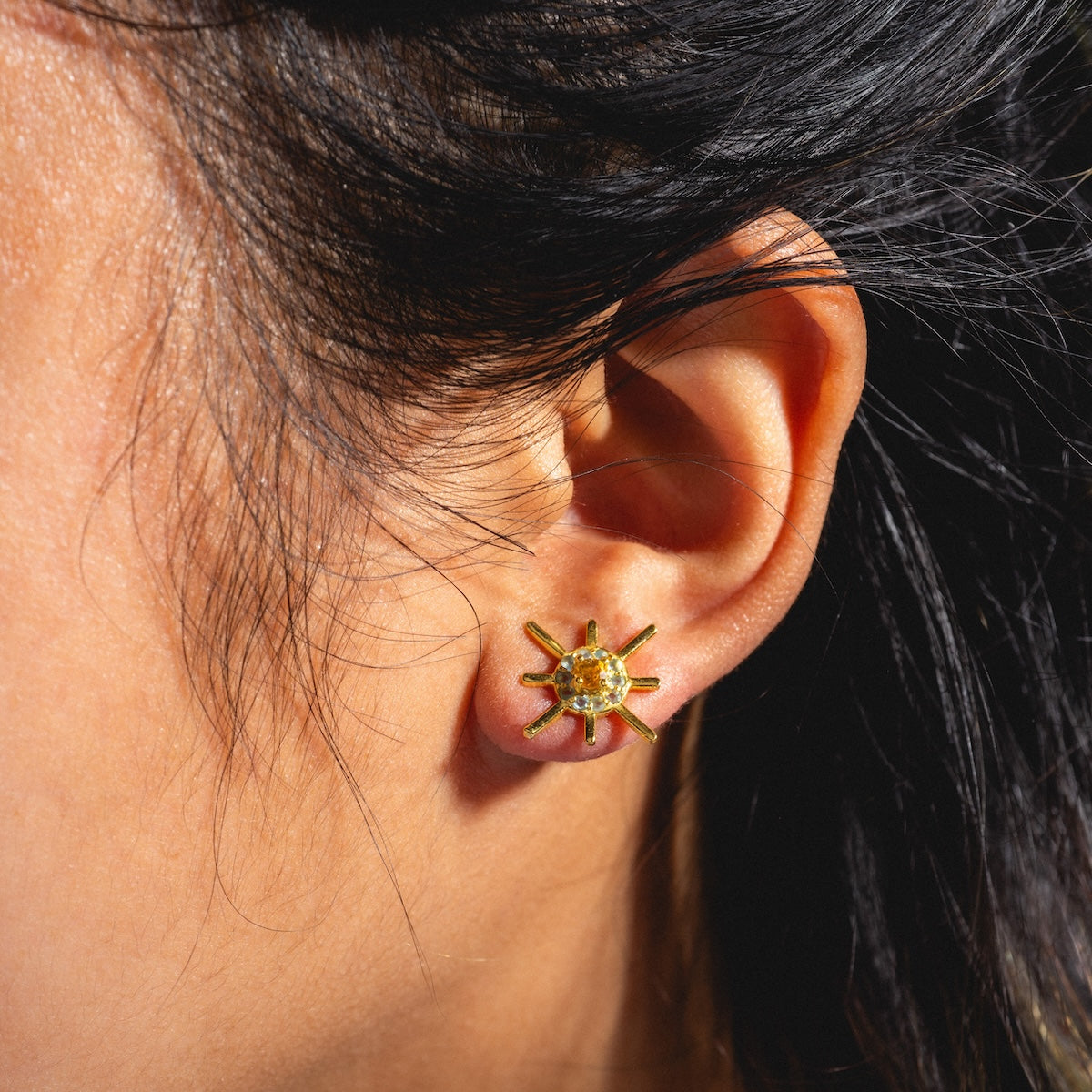 Close-up of a persons ear wearing Sol Stud Earrings shaped like a sun, with a yellow gemstone center. Their dark hair is tucked behind the ear.