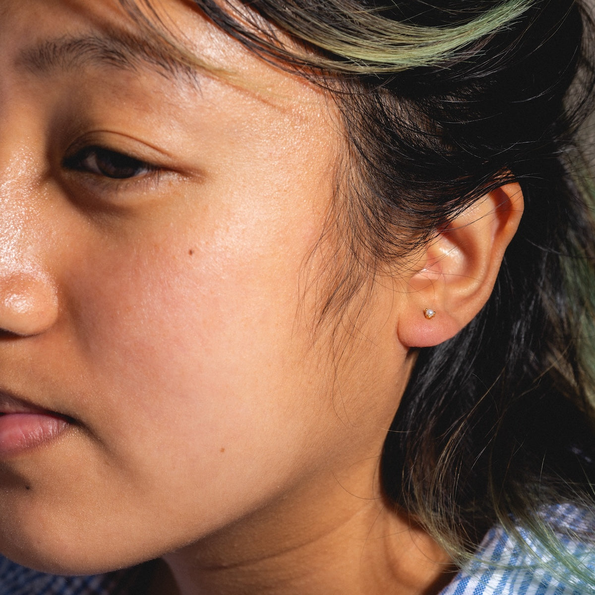 Close-up of a calm person with light brown skin, straight dark hair with green highlights, and wearing Fabulous Studs—small 14k gold plated earrings—while looking slightly down.