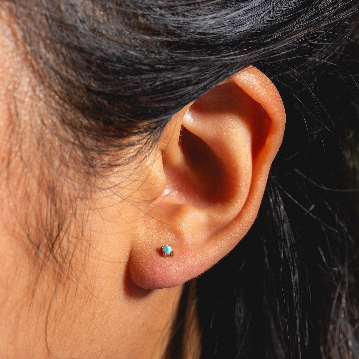 Close-up of a person’s ear wearing Fabulous Studs—a small opal stud earring set in 14k gold plating, accentuating the turquoise tone. The wearer has dark hair and smooth, well-lit skin.