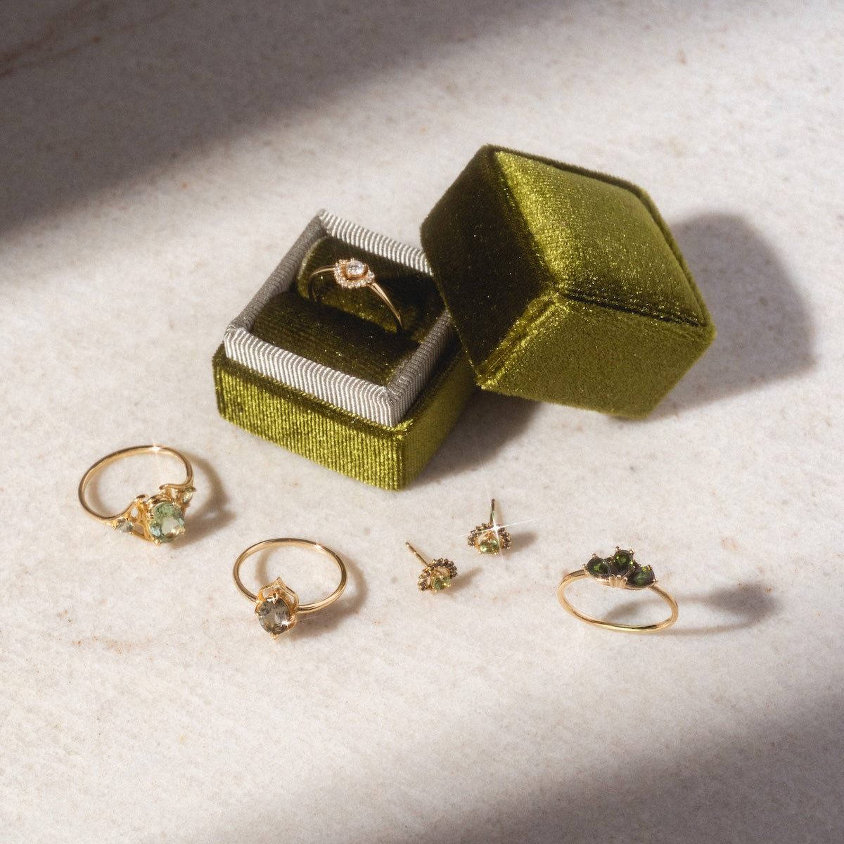 Tosia Tourmaline Studs and other gold jewelry, including rings and earrings, are arranged on a light surface. An olive green velvet ring box holds a 14k gold ring with a clear stone. Shadows fall across the scene.