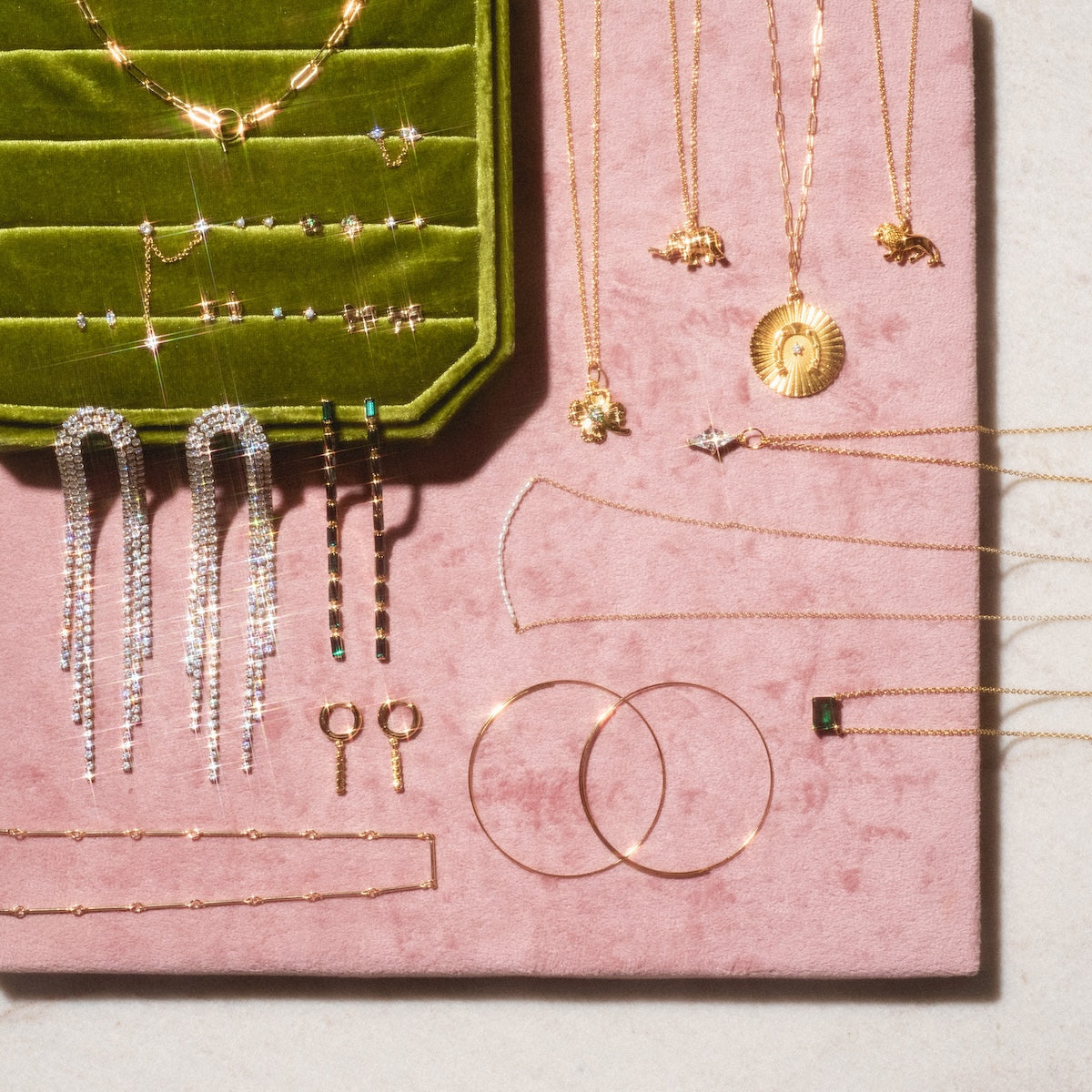 The Wise Necklace, a 14k gold plated elephant pendant, is displayed with assorted gold and silver rings, bracelets, earrings, and studs on green velvet and pink suede trays in bright natural light.