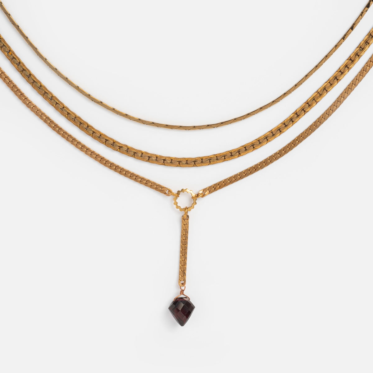 The Palmer Gemstone Necklace is a three-layered gold piece made from recycled brass chains, featuring unique styles with a small circular detail and a dark red garnet pendant on the lowest chain, displayed against a white background.