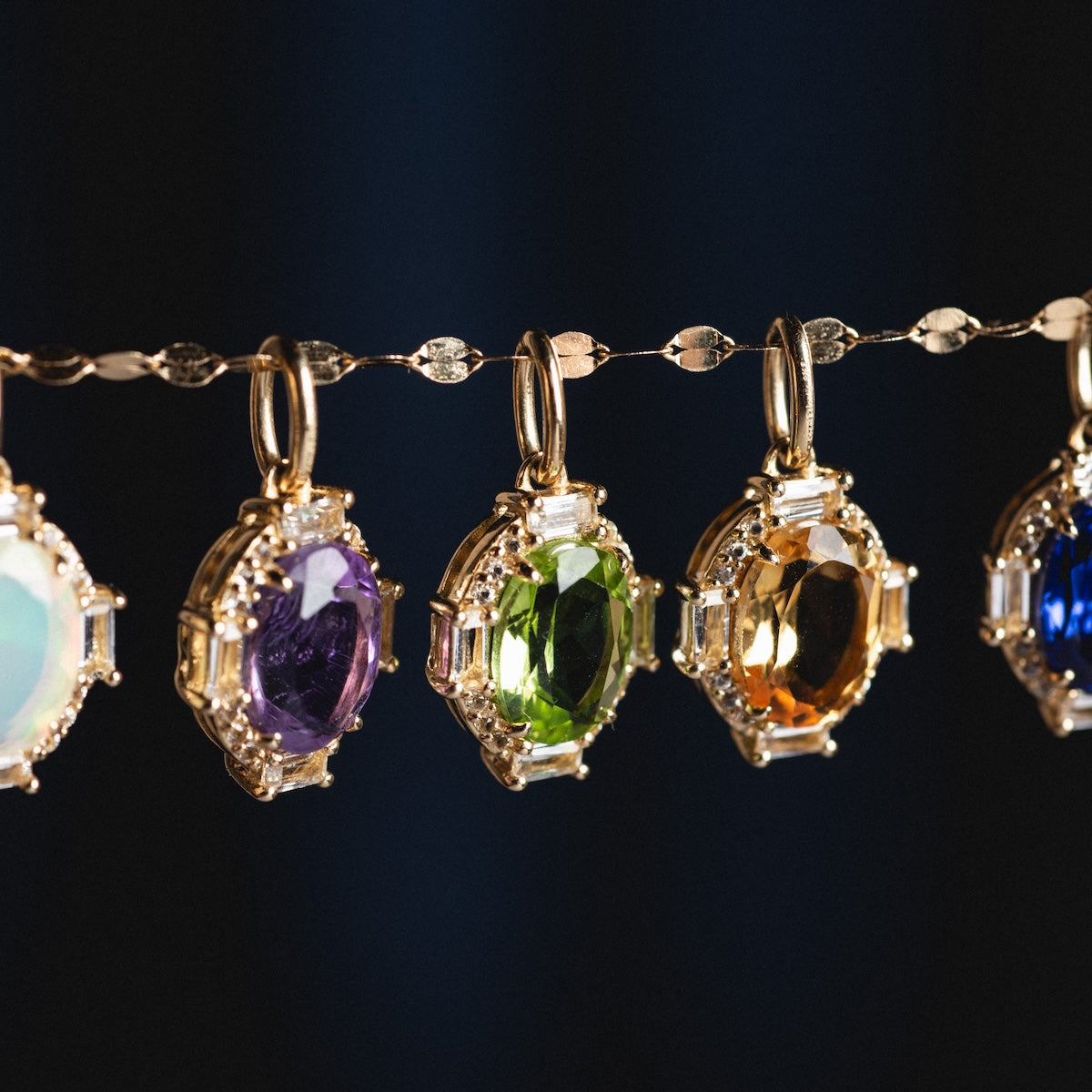 Five Solid Gold Vintage Inspired Birthstone Charms with oval white, purple, green, orange, and blue gemstones hang from a decorative gold chain against a dark background.