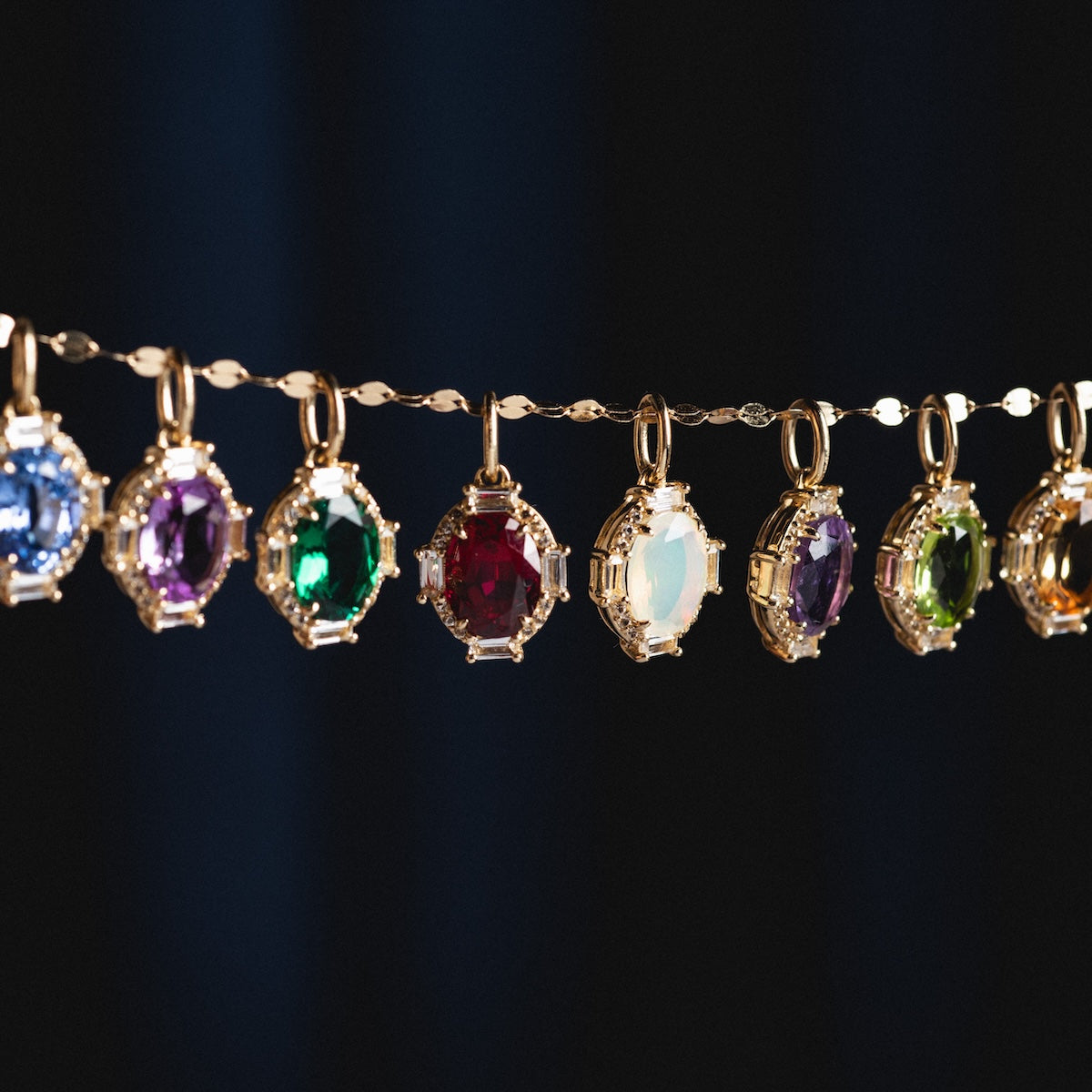 A Solid Gold Vintage Inspired Birthstone Charm with colorful stones—blue, purple, green, red, milky white, and amber—hangs from a delicate gold chain against a dark background.