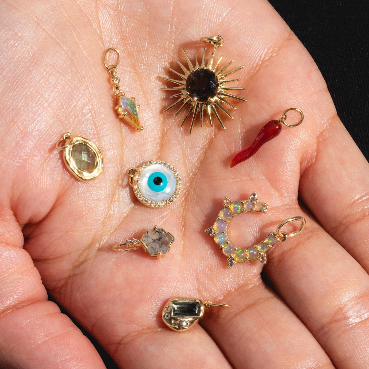 A hand holds the Solid Gold Amulet Charm set—eight small, colorful pieces including a sunburst, evil eye, red horn, crescent moon with stones, faceted gems, and a gold-framed translucent pendant—ideal for unique gemstone necklaces.