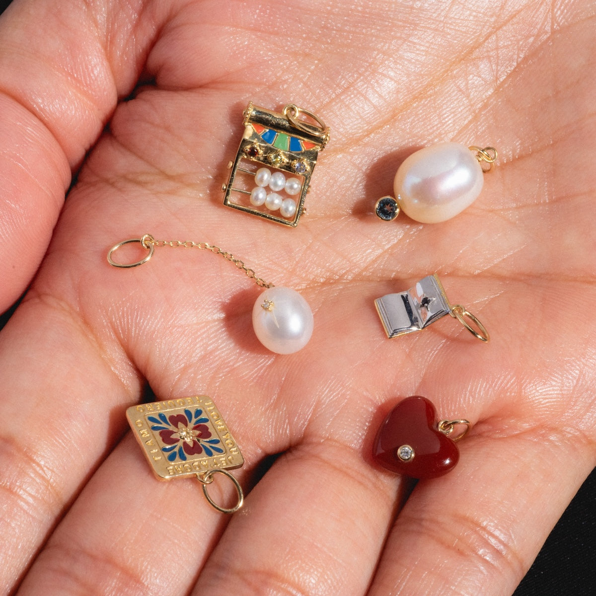 A hand holds the Solid Gold Statement Charm, a 9k yellow gold book with pearls, along with a gold chain featuring a pearl, an enamel rectangle charm, a silver book, and a red heart charm adorned with a small gemstone.