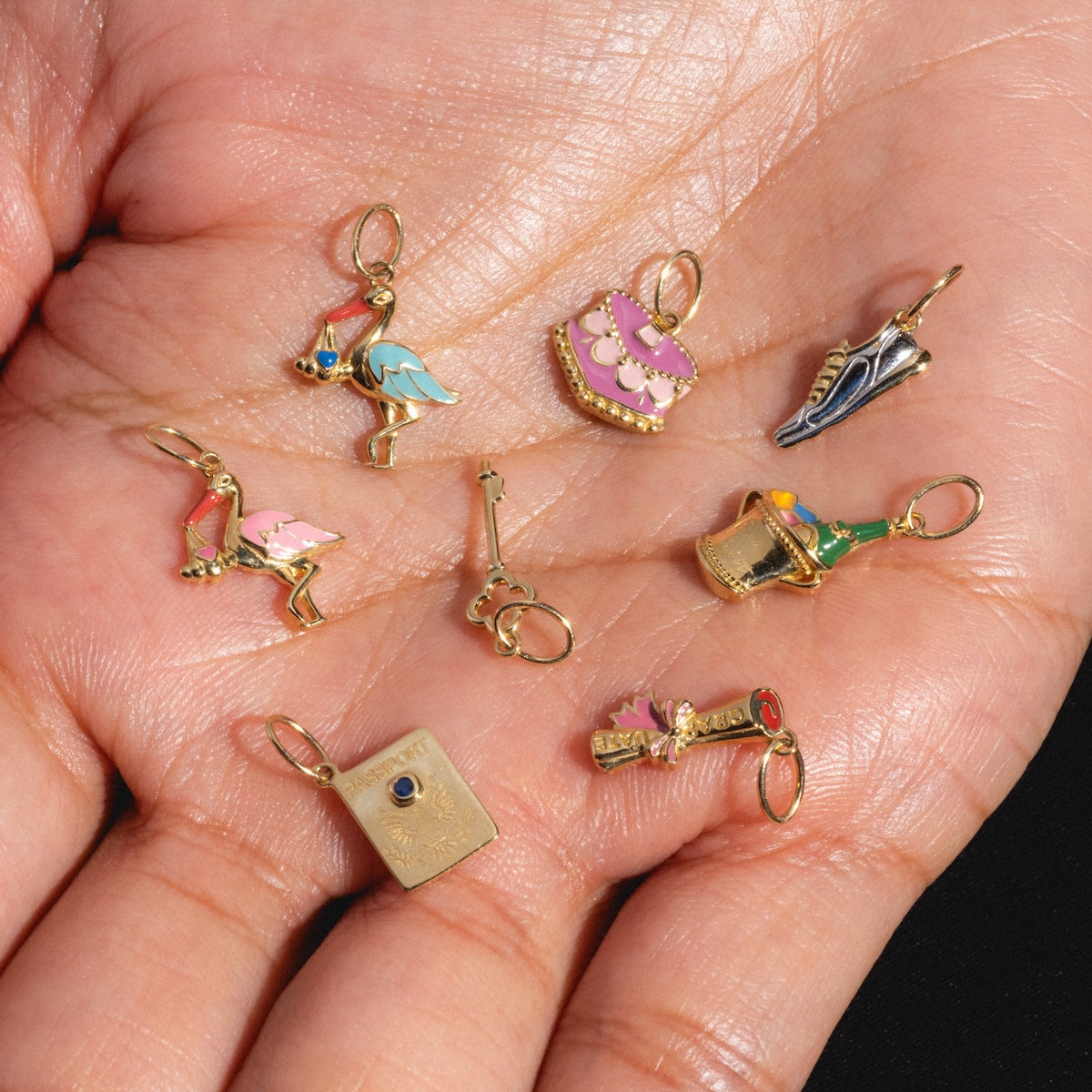 A hand holds eight Solid Gold Celebration Charms—miniature book, shoe, crown, bottle, flamingo, teapot, dress, and key—each with gold accents or enamel details, perfect as engraved gold charms for a unique touch.