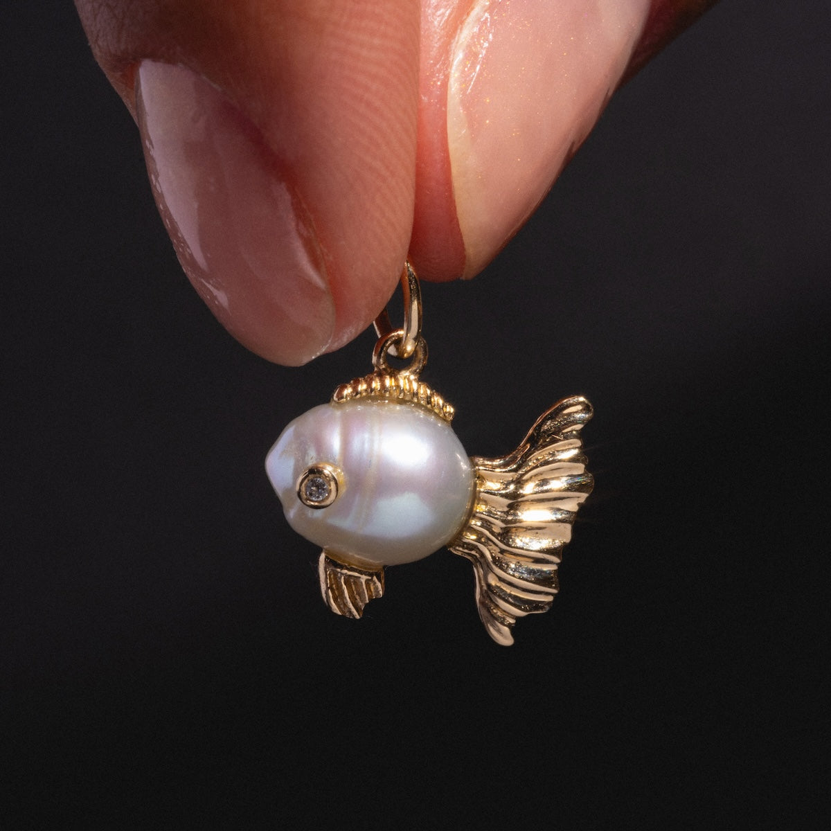A hand holds a Solid Gold Nature Charm—a fish-shaped gold pendant with a round pearl body, detailed gold fins and tail, and a small gemstone eye—shown against a dark background.