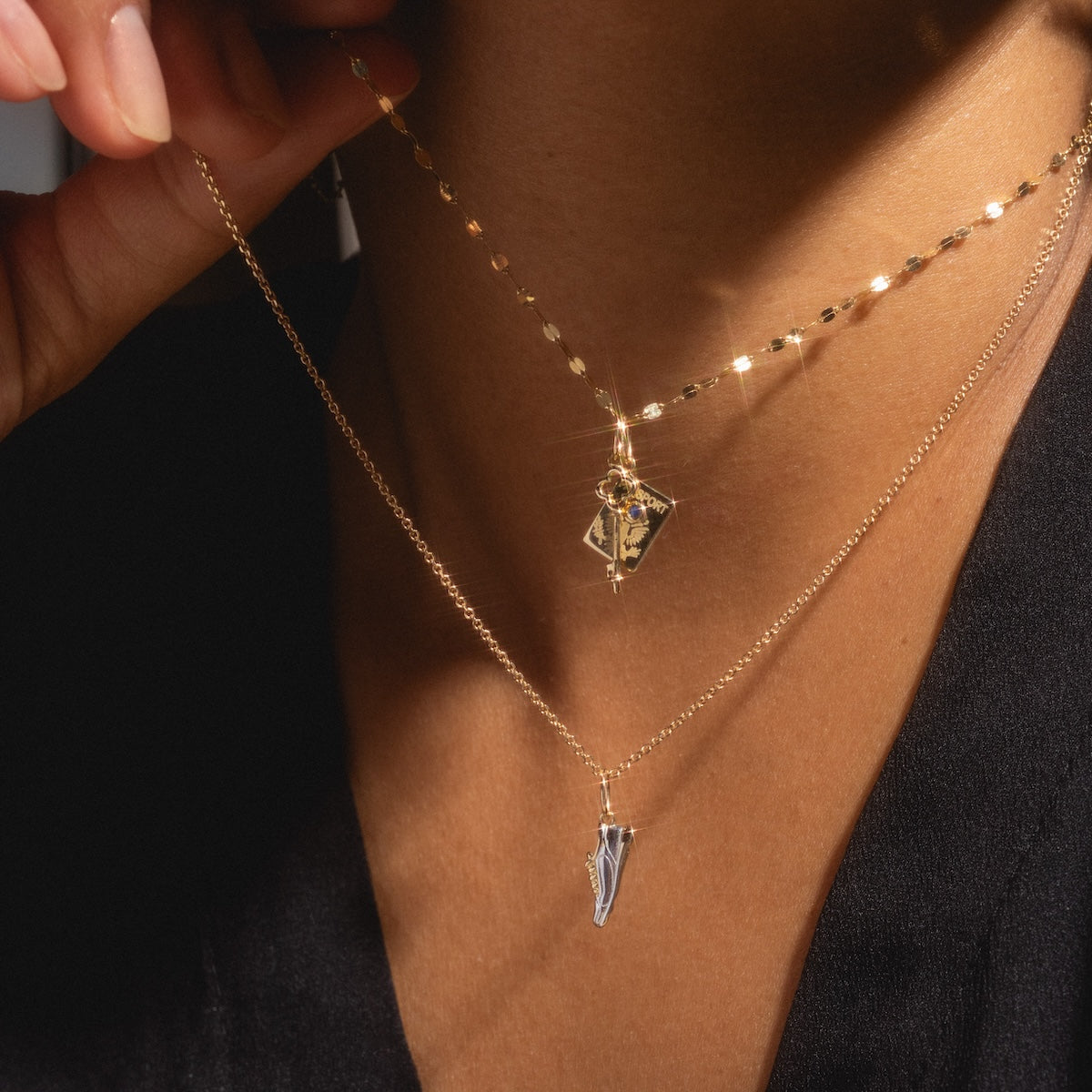 A close-up of a person wearing the Solid Gold Celebration Charm, featuring two 9k solid yellow gold necklaces—one with a geometric gemstone pendant and another with a sleek, pointed charm. The person gently holds one necklace.