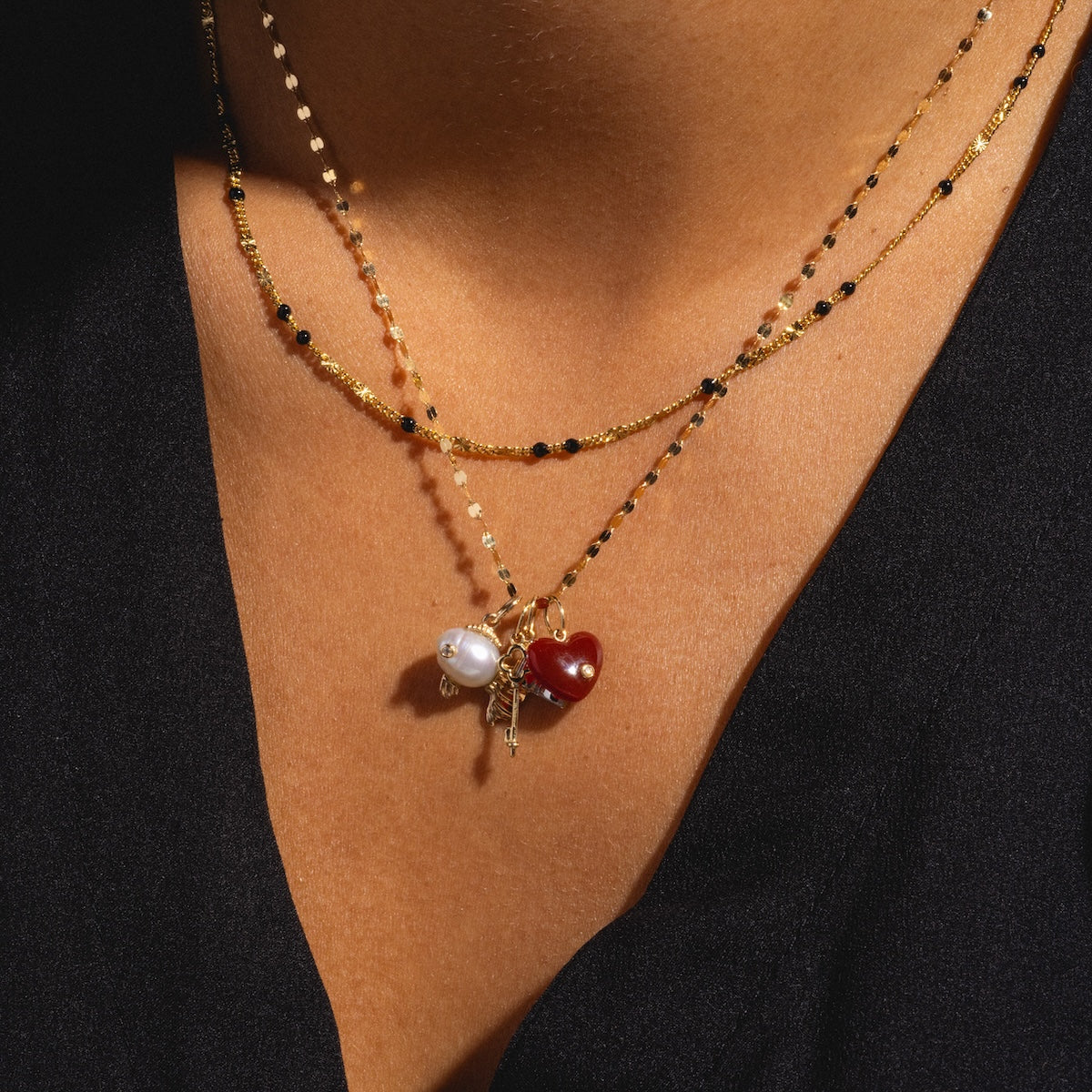 A close-up of a person wearing two gold necklaces—one with a white pearl and blue topaz pendant, the other featuring the Solid Gold Statement Charm—styled over a black V-neck top.