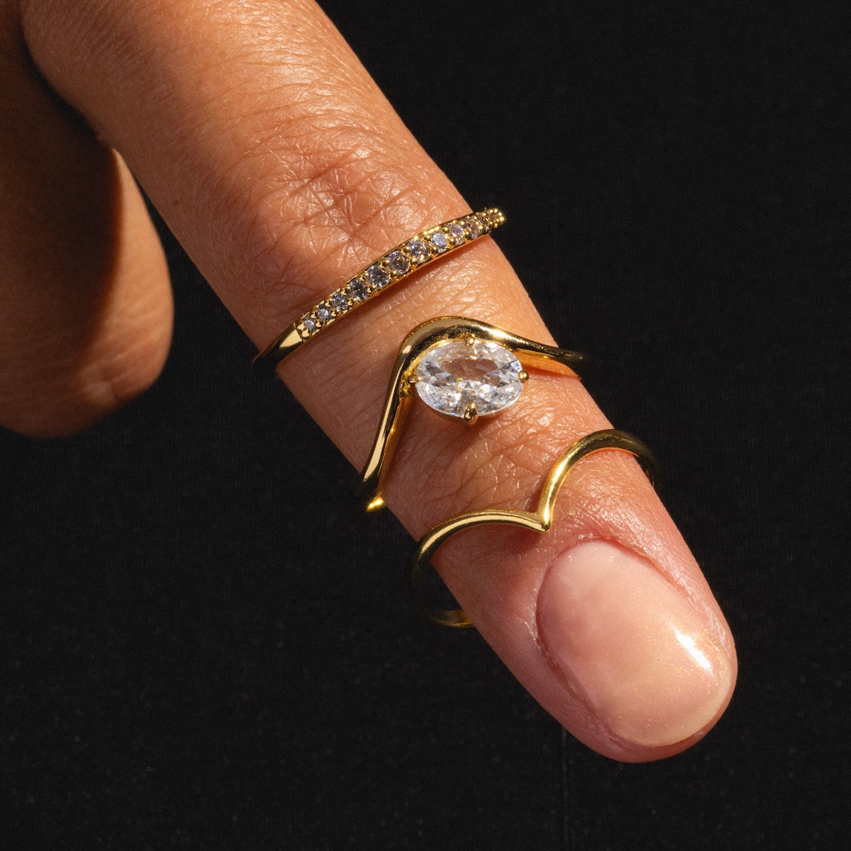 A close-up of a hand with three gold rings on the index finger, including the Arch Ring—an 18k yellow gold plated stacking ring with small diamonds—a wavy minimalist ring, and one with a large clear gemstone, set against a black background.
