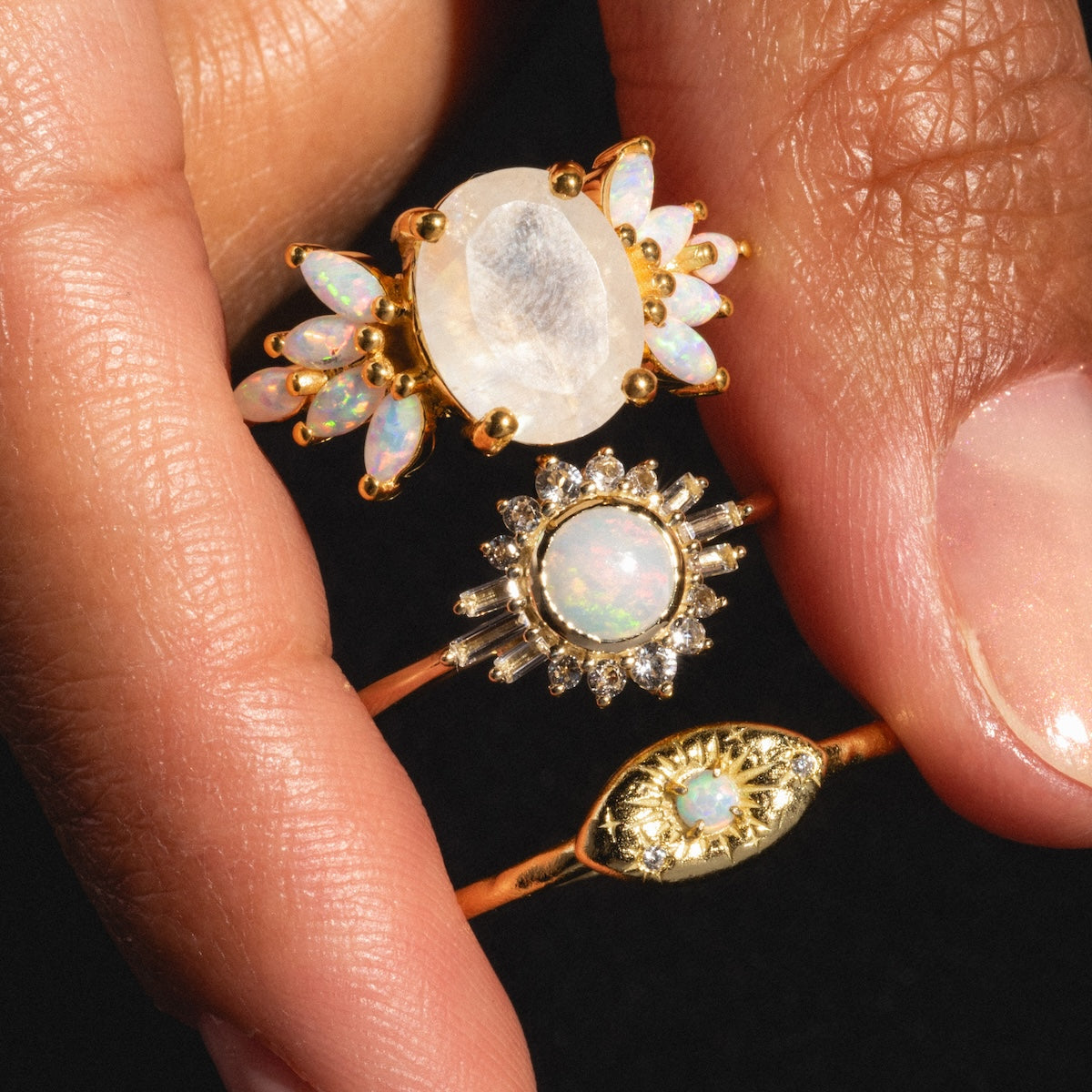 A close-up of three gold rings on a persons fingers, including the Opal Amulet Ring with intricate designs and sparkling stones, set against a black background.