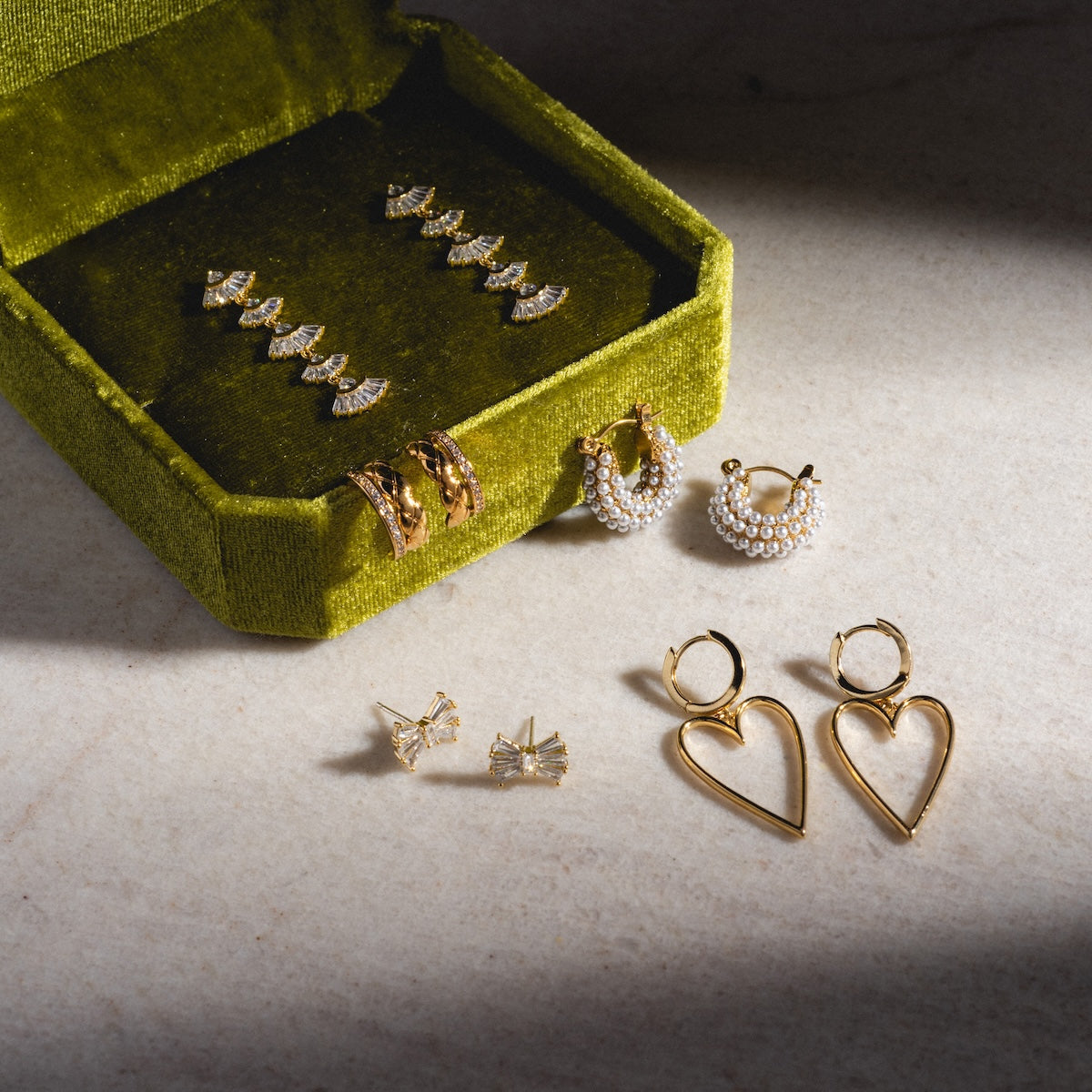 A green velvet jewelry box displays the Rene Drop Earrings—gold and pearl art deco-inspired drops—arranged elegantly alongside geometric studs, hoops, pearl hoops, and large heart-shaped dangles on marble.