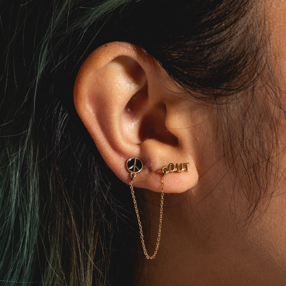 A close-up of an ear wearing the Peace Out Double Pierce Earring Sample: two 18k yellow gold plated earrings—one with a peace sign, one spelling OUT—connected by a delicate chain. The wearer’s dark hair has hints of green.