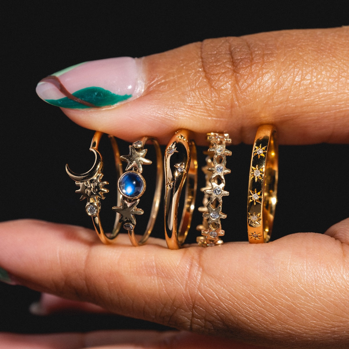 A hand with green and pink nail art displays the Cosmic Dome Ring, a gold plated piece featuring celestial designs like stars, a crescent moon, gemstones, and intricate patterns, set against a black background.