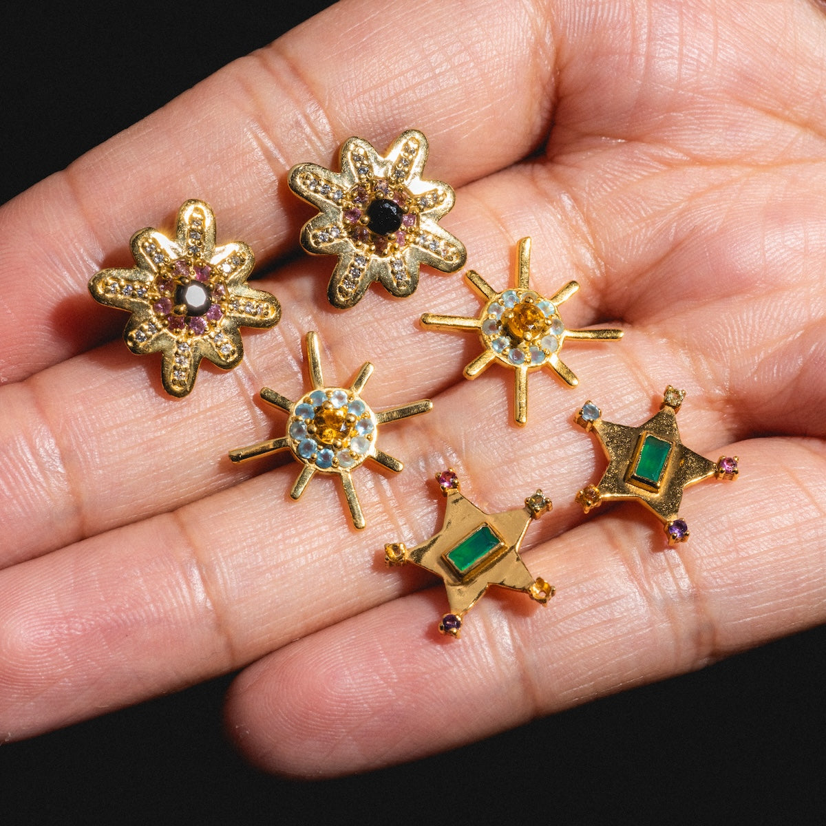 A hand holds three pairs of gold earrings: floral with a dark center, star-shaped with green and colorful gems, and sun-shaped Sol Stud Earrings adorned with citrine and blue chalcedony stones.