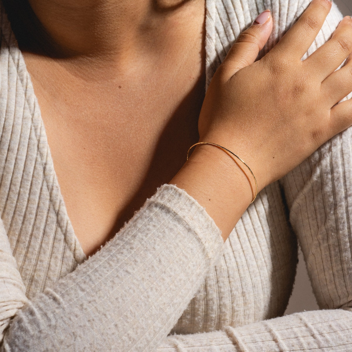 A person wearing a textured cream long-sleeve top rests their hand on their shoulder, showcasing the Solid Gold Structured Bracelet crafted from 9k solid gold on their wrist.