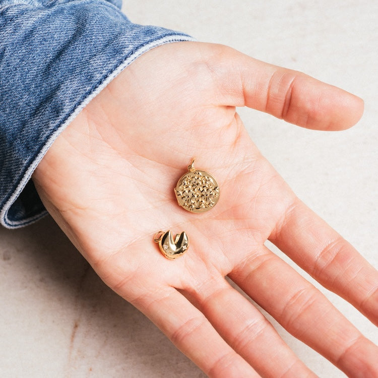 A hand in a denim sleeve holds two gold pendants, including the Solid Gold Locket Charm shaped like a stylized “V” or heart, alongside another with a raised geometric pattern.