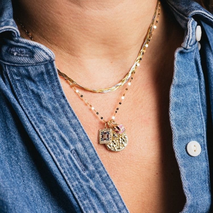 A person wears a blue denim shirt with three layered gold necklaces, including a twisted chain, a beaded pendant chain, and one featuring the Solid Gold Locket Charm alongside a round textured coin.