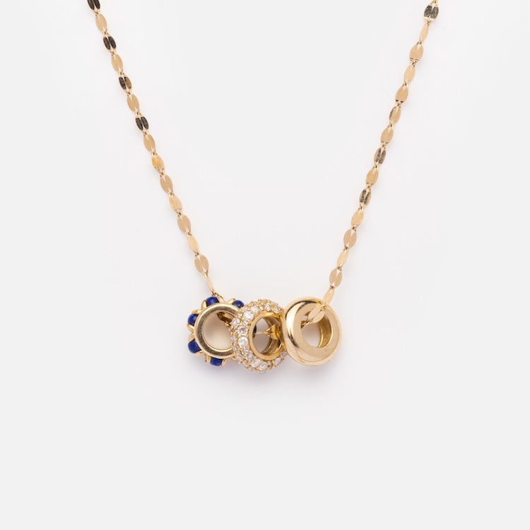 The Solid Gold Anniversaire Charm is a gold chain necklace with three circular pendants—one with blue enamel, one with clear crystals, and one plain gold—perfect for celebrating life’s milestones.