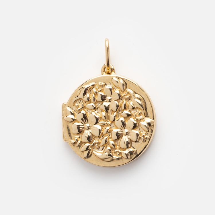 The Solid Gold Locket Charm is a round locket with a raised floral design, showcasing detailed flowers and leaves on the front, set against a plain white background.