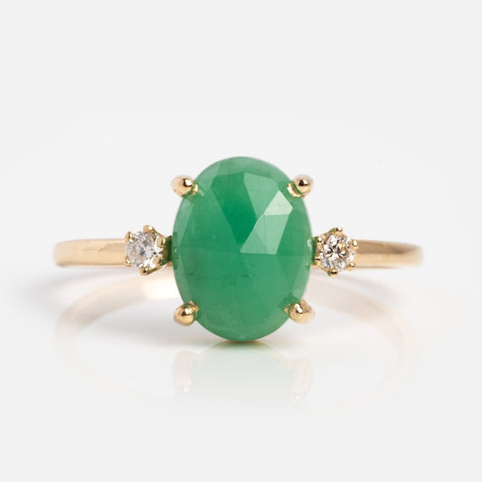 The Oval Emerald Ring with Diamonds in Yellow Gold features a large oval green gemstone at the center, accented by two round diamonds on each side, and is displayed against a plain white background.