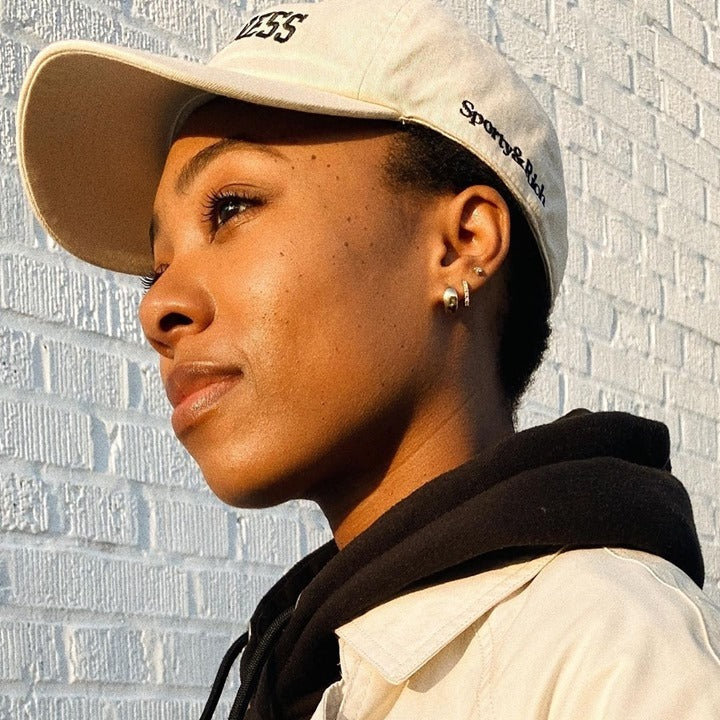 A person in a beige cap, black hoodie, and Solid Gold Chunky Huggie Hoops stands outside by a white brick wall, sunlight illuminating their face as they gaze thoughtfully into the distance.