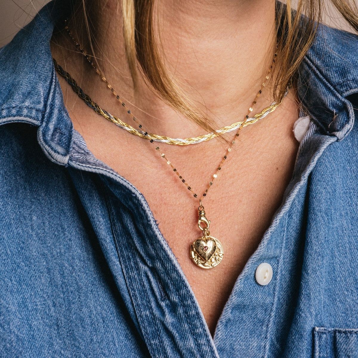 A person wearing a blue denim shirt and two gold necklaces, one featuring the Solid Gold Locket Charm—a heart-shaped locket with a floral etched design.