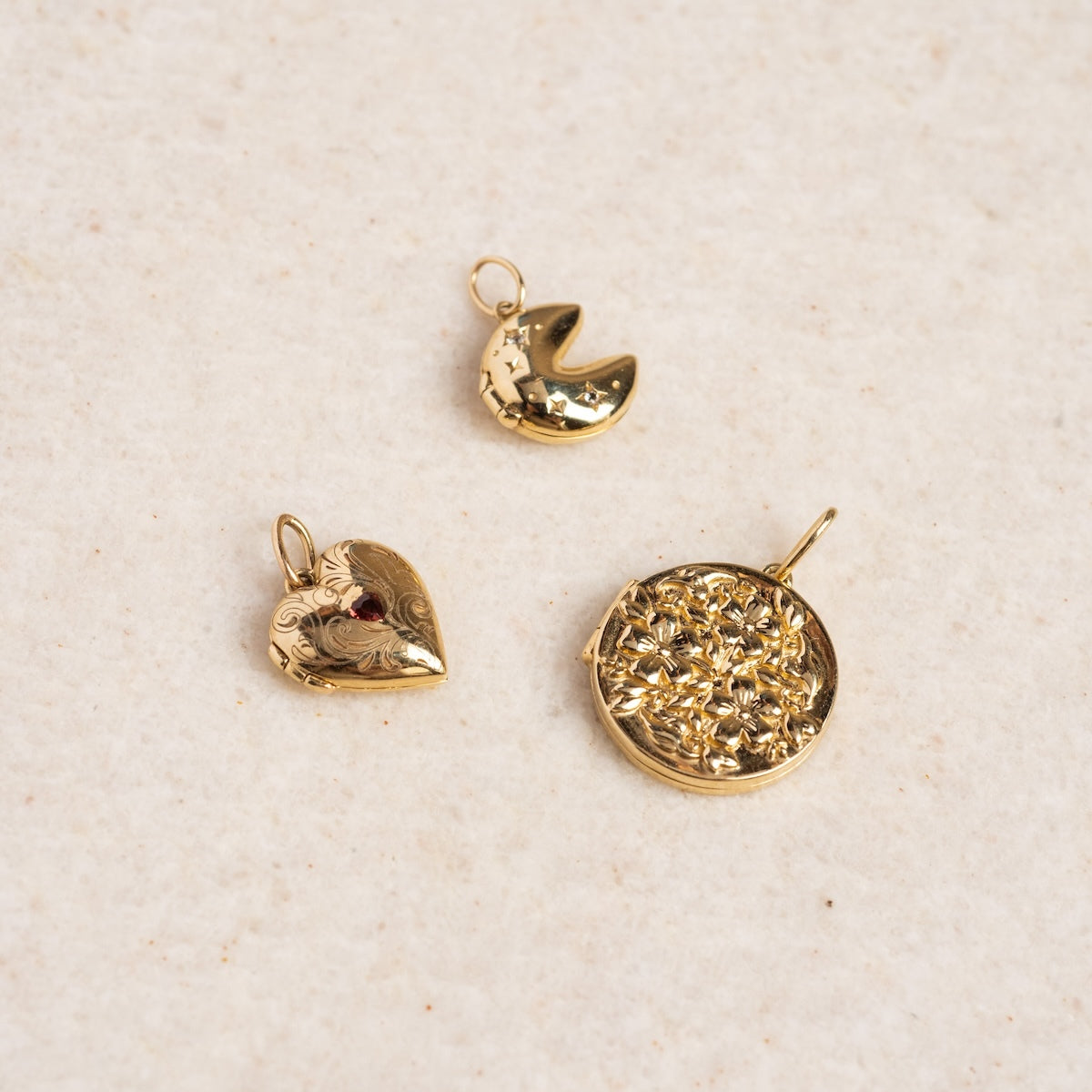 A Solid Gold Locket Charm, a small crescent moon pendant, and a round floral-patterned pendant are displayed on a lightly textured surface.
