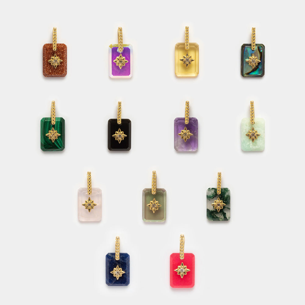 Twelve Solid Gold Crystal Amulet Charms for Manifestation, each in a unique color and pattern with gold star accents and textured gold bails, are arranged in a neat grid on a white background.