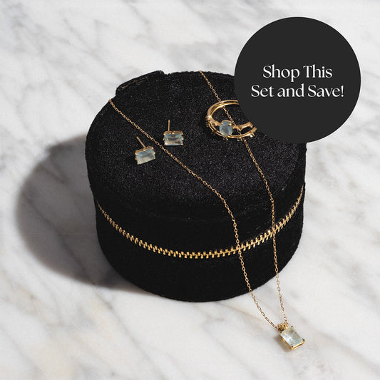 A velvet black box displays the Solid Gold 2025 March Capsule Aquamarine Full Set—necklace, earrings, and ring. A dark circle overlay says, “Shop This Set and Save!” All shown on a white marble background. Free gift included.