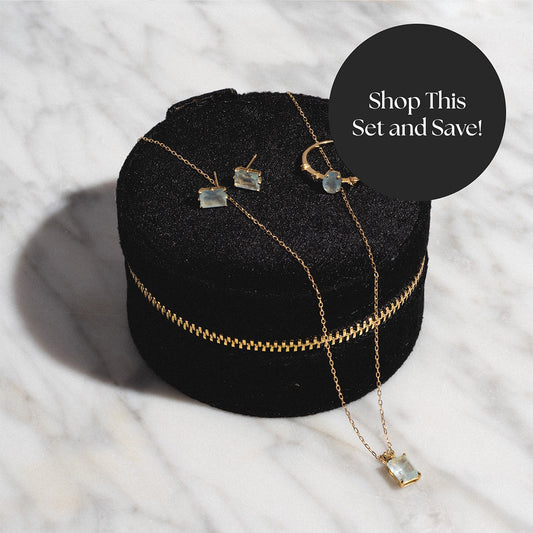 The Solid Gold 2025 March Capsule Aquamarine Statement Set with Free Gift, featuring a necklace, ring, and earrings with light blue aquamarine stones, is displayed on a round black velvet box labeled Shop This Set and Save! on a marble surface.
