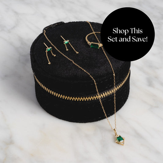 The Solid Gold 2025 May Capsule Emerald Statement Set, featuring earrings, bracelet, and necklace, is elegantly displayed on a black velvet case. A black circle reads: Shop This Set & Save! Includes Free Gift—perfect May birthday present.