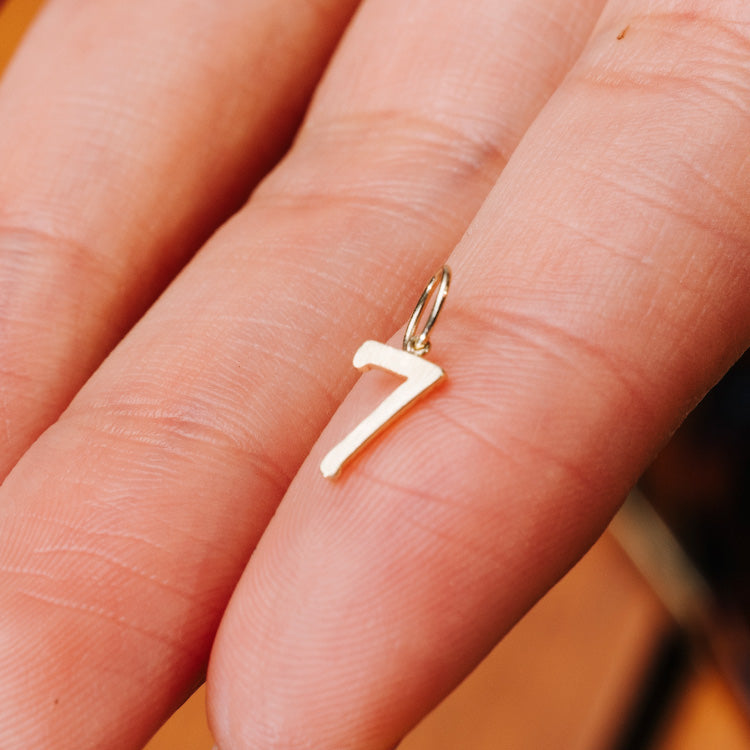 A close-up of a hand holding a Solid Gold Number Charm shaped like the number 7, featuring a small loop at the top—ideal for attaching to a chain or bracelet and adding to your personalized jewelry collection.